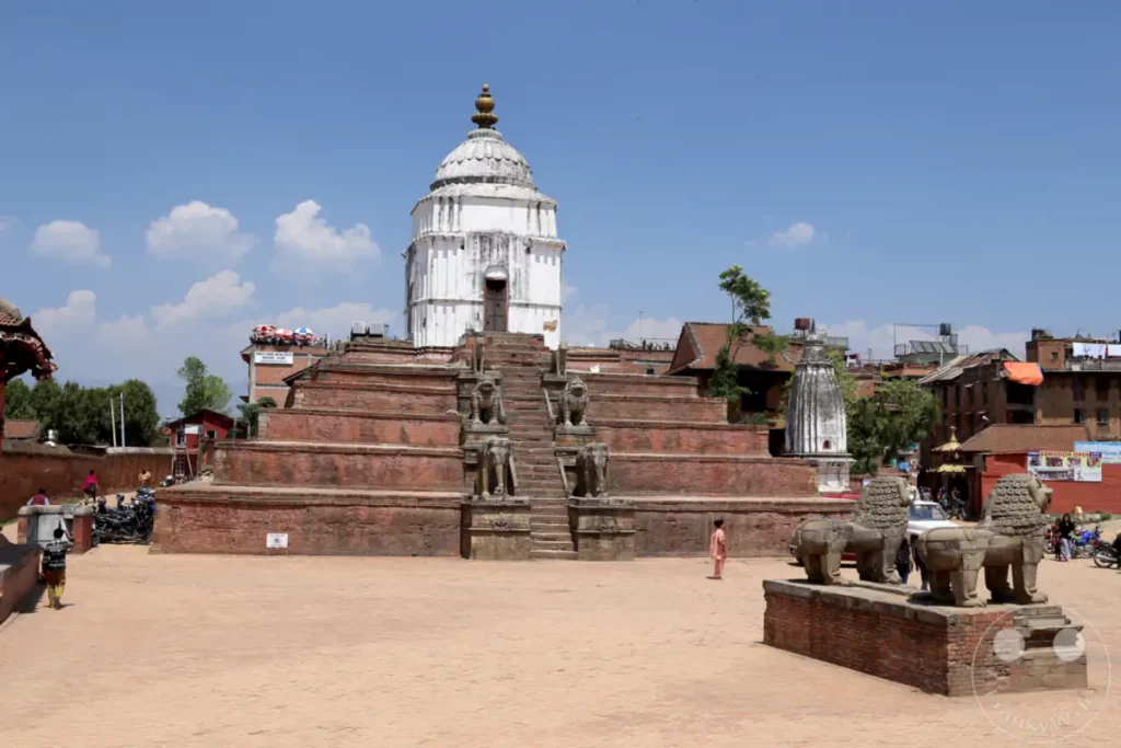 Ein weitläufiger Platz in Bhaktapur, Nepal, mit einem markanten Stufentempel, der von steinernen Löwenstatuen flankiert wird. Im Hintergrund sind weitere traditionelle Gebäude und ein weißer Tempel zu sehen, was auf eine historische und kulturelle Stätte hindeutet.