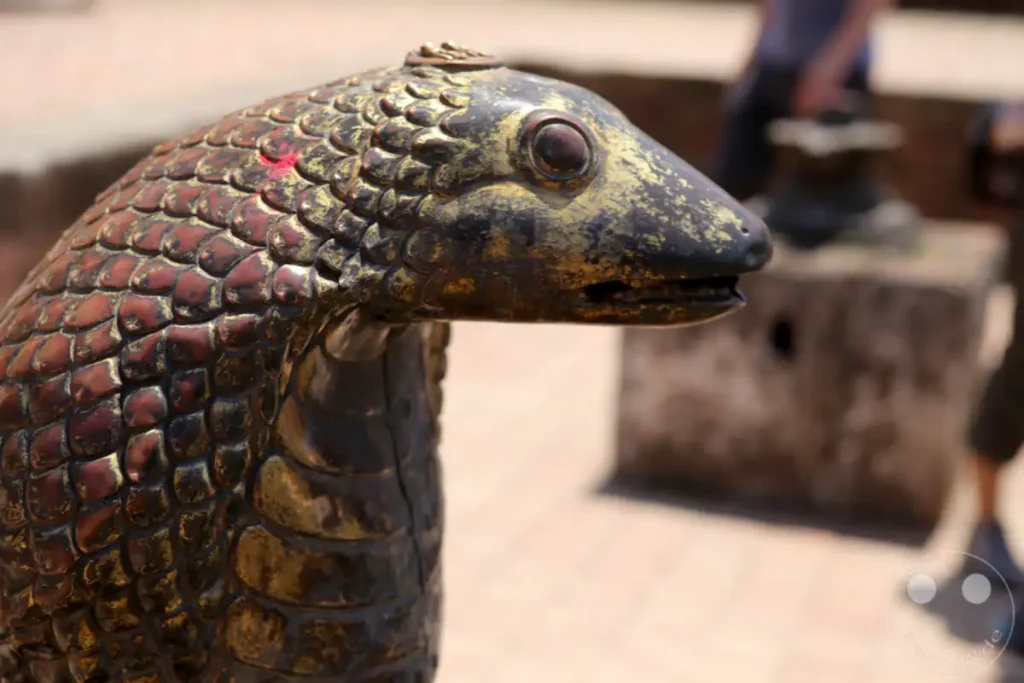 Nahaufnahme einer detailreich verzierten Tierfigur aus Metall in Bhaktapur, Nepal. Die Figur ist mit schuppenähnlichen Mustern und goldenen Akzenten verziert und zeigt feine Details wie das Auge und den Mund. Im Hintergrund verschwimmen unscharf Personen und Bauwerke, was die Fokussierung auf das Kunstwerk verstärkt.