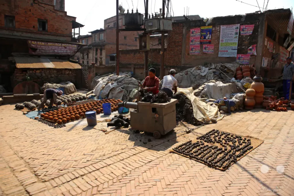 In Bhaktapur, Nepal, trocknen mehrere Personen unfertige Töpferwaren auf einem mit Ziegeln gepflasterten Hof. Die Szene zeigt den traditionellen Herstellungsprozess von Keramikprodukten in einem ländlichen Umfeld unter freiem Himmel.