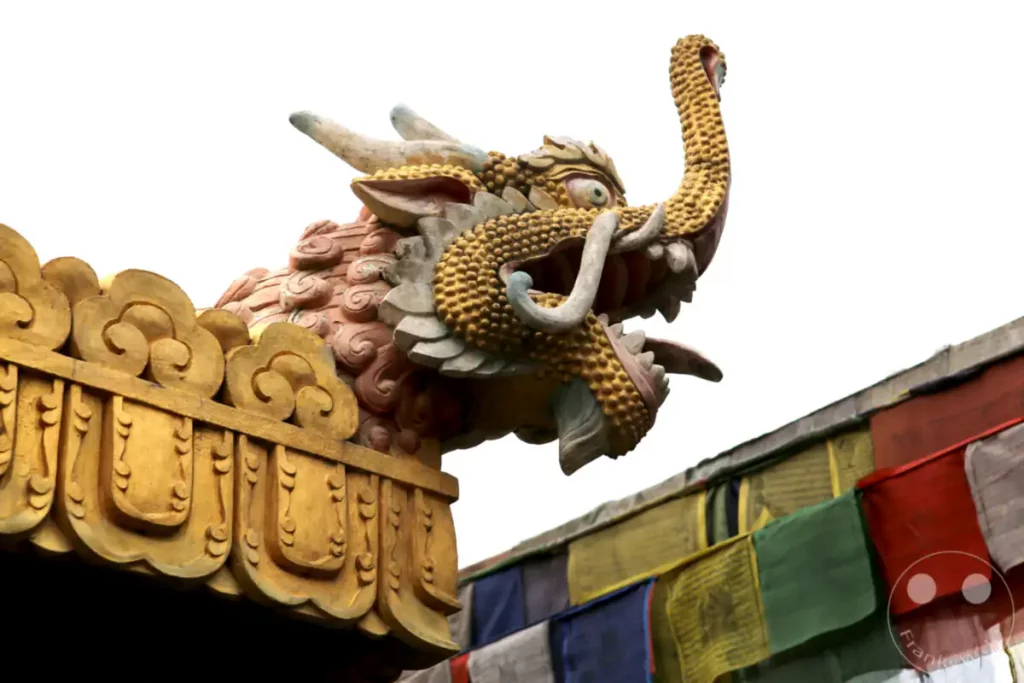 Detailaufnahme einer kunstvollen Drachenkopf-Skulptur auf dem Dach der Boudhanath-Stupa in Nepal. Bunte Gebetsfahnen im Vordergrund verleihen dem Bild eine lebendige und spirituelle Atmosphäre, die die kulturelle Bedeutung des Ortes unterstreicht.