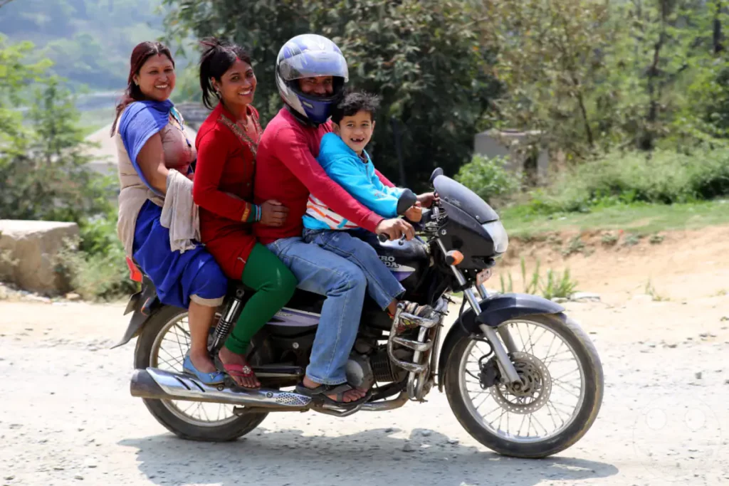 Eine Familie bestehend aus zwei Frauen und einem Kind fährt gemeinsam auf einem Motorrad über eine staubige Straße in der Nähe der Chandra Bridge in Nepal. Alle lächeln und scheinen die gemeinsame Fahrt zu genießen, trotz der ungewöhnlichen Beladung des Fahrzeugs.