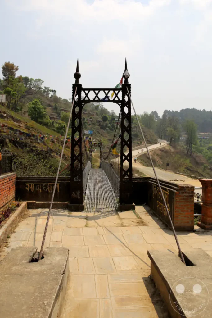 Eine robuste Hängebrücke überspannt ein Tal und ist mit bunten Gebetsfahnen geschmückt. Im Hintergrund erstreckt sich eine grüne Berglandschaft mit kleinen Siedlungen und einer kurvenreichen Straße, die zum Wandern einlädt. Die Szene strahlt eine friedliche Atmosphäre aus und vermittelt ein Gefühl von Abenteuer und Spiritualität.