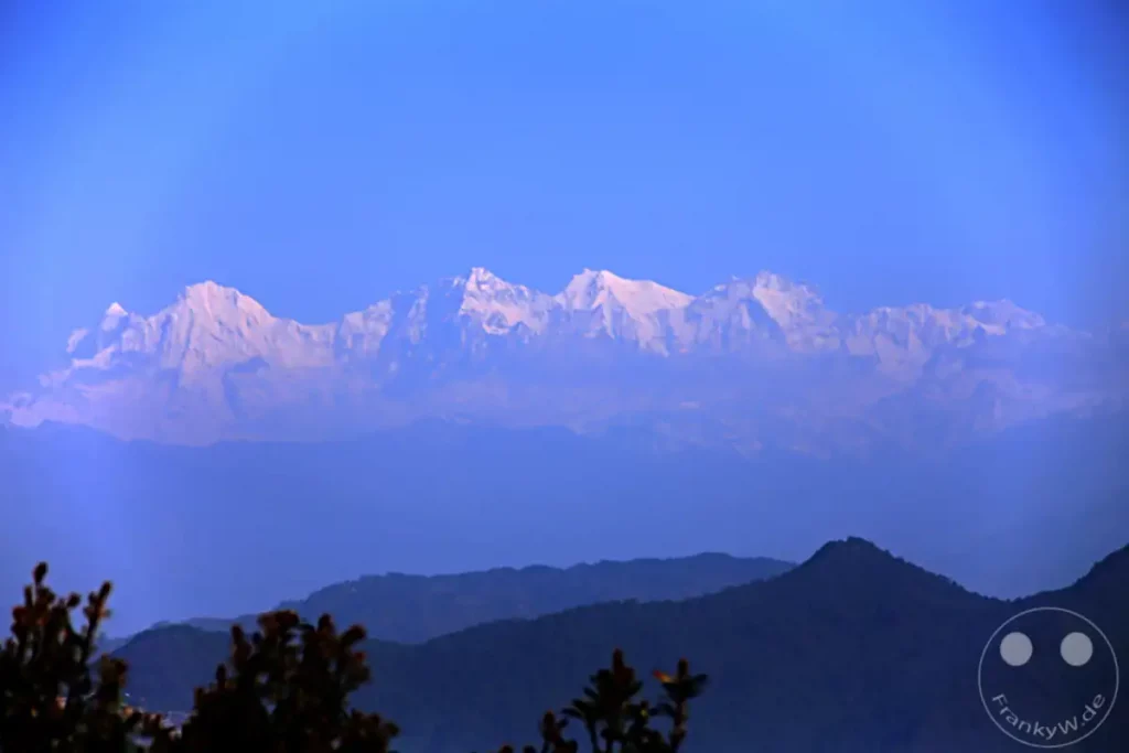 Panorama von schneebedeckten Berggipfeln des Himalaya, die sich im Dunst vor einem klaren, blauen Himmel abzeichnen. Die Szene, aufgenommen in Dakshinkali, vermittelt eine friedliche und majestätische Atmosphäre in der blauen Stunde.