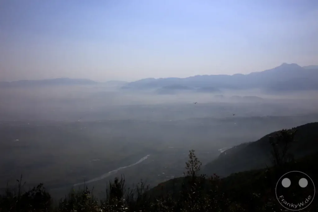 Die weite Berglandschaft des Himalaya-Gebirges liegt in Morgennebel gehüllt, während die ersten Sonnenstrahlen die Gipfel küssen. Das Tal unterhalb wird von einem Fluss durchzogen, während vereinzelte Lichter einer Siedlung durch den Dunst schimmern.