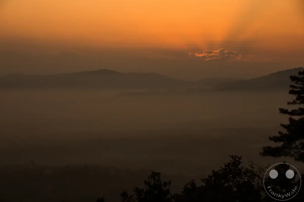 Ein dramatischer Sonnenuntergang über den Bergen von Dakshinkali. Die letzten Strahlen der Sonne brechen durch eine einzelne Wolke und beleuchten die nebelverhangenen Täler. Die Silhouette des Himalaya-Gebirges und die dunklen Bäume im Vordergrund erzeugen eine mysteriöse und friedliche Atmosphäre.