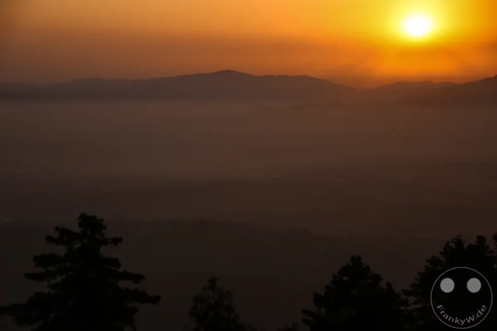 Der dramatische Sonnenuntergang taucht die Berglandschaft von Dakshinkali in ein warmes, orangefarbenes Licht. Der aufsteigende Dunst verschleiert die darunterliegende Ebene und lässt die Berge des Himalaya-Gebirges und die Silhouetten der Bäume in der Dunkelheit verschwinden.