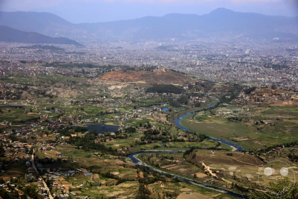 Diese Aufnahme von Dakshinkali bietet einen weiten Blick über eine dicht besiedelte Stadt, die von sanften Hügeln und landwirtschaftlich genutzten Flächen umgeben ist. Ein Fluss schlängelt sich durch die Szenerie und verbindet die städtische Ausdehnung mit der umliegenden Natur. Die Atmosphäre ist ruhig und erhaben, trotz der urbanen Präsenz im Hintergrund.