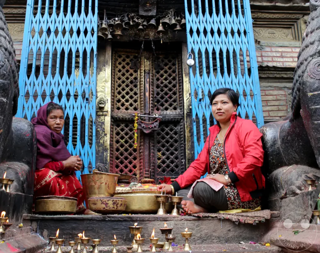 Zwei Frauen sitzen vor dem Eingangsportal des Aanandadi Lokeshwar Tempels in Kathmandu, Nepal. Sie sind von zahlreichen Opfergaben und brennenden Öllampen umgeben, die eine feierliche Atmosphäre schaffen. Die Szene spiegelt tiefen Glauben und religiöse Rituale im Tempel wider.