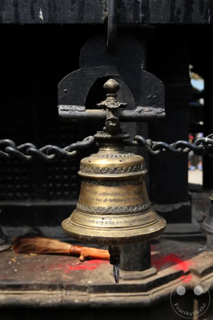 Eine kunstvoll verzierte Glocke aus Messing hängt an einer schweren Kette vor dem Anandadi Lokeshwar Tempel in Kathmandu. Inschriften in nepalesischer Schrift zieren die Oberfläche der Glocke, während im Hintergrund eine traditionelle Besen und rote Farbe auf den Stufen sichtbar sind.