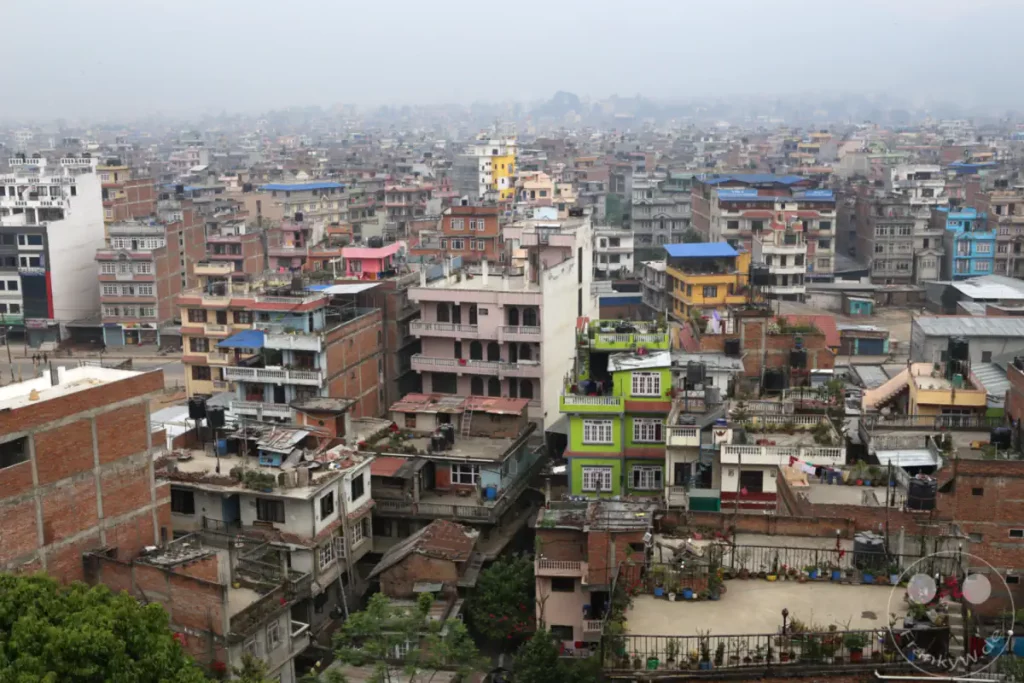 Die Aufnahme bietet einen weiten Blick über die dicht bebaute Stadt Kathmandu, Nepal. Unzählige Gebäude unterschiedlichster Größe und Bauweise sind zu sehen, die meisten mit flachen Dächern und vielen Wasserbehältern. Eine Dunstschicht liegt über der Stadt und vermittelt eine gedämpfte, alltägliche Atmosphäre.