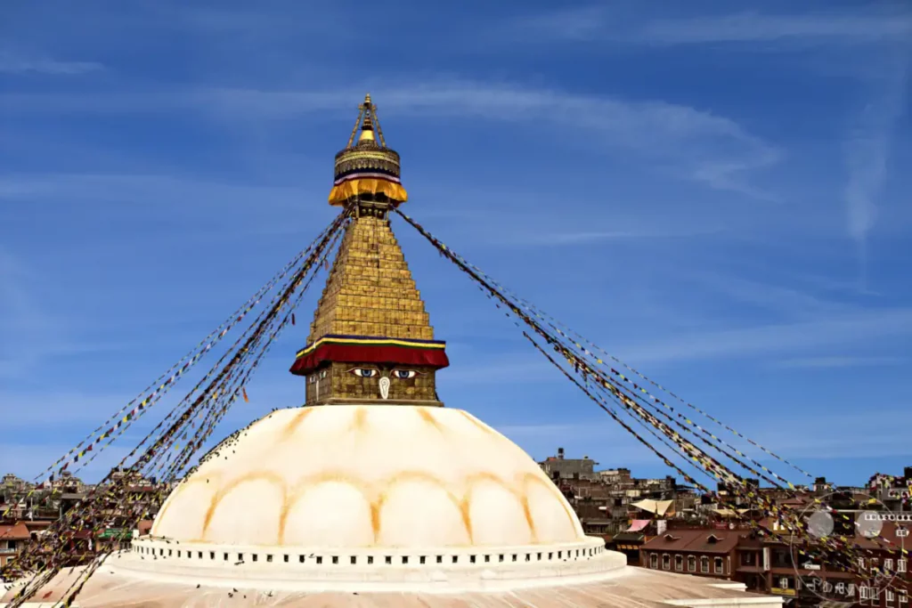 Die monumentale Boudhanath-Stupa in Kathmandu unter einem strahlend blauen Himmel mit feinen Schleierwolken. Die weiße Kuppel bildet die Basis für den goldenen Turm, auf dem die charakteristischen Augen Buddhas und bunte Gebetsfahnen zu sehen sind. Die Szenerie vermittelt eine friedliche, spirituelle und zugleich majestätische Atmosphäre.