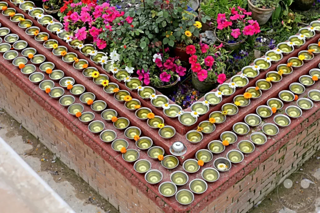 Am Rand der Boudhanath-Stupa in Kathmandu sind Reihen von silbernen Schalen mit Wasser und Blumen für religiöse Rituale aufgestellt. Die Dekoration mit bunten Blüten und Pflanzen unterstreicht die spirituelle Atmosphäre des buddhistischen Heiligtums.