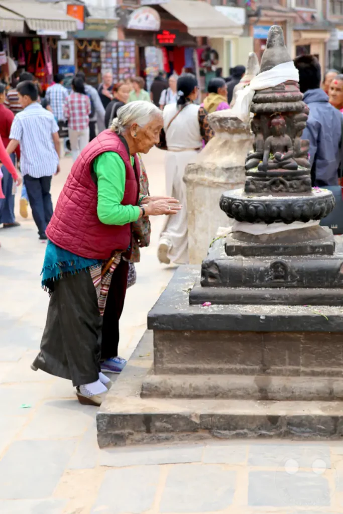 Eine ältere Pilgerin verrichtet in Anbetracht eines buddhistischen Schreins an der Boudhanath-Stupa in Kathmandu ihre Andacht. Die Szene spielt sich in einer belebten Umgebung mit anderen Menschen und Marktständen ab.