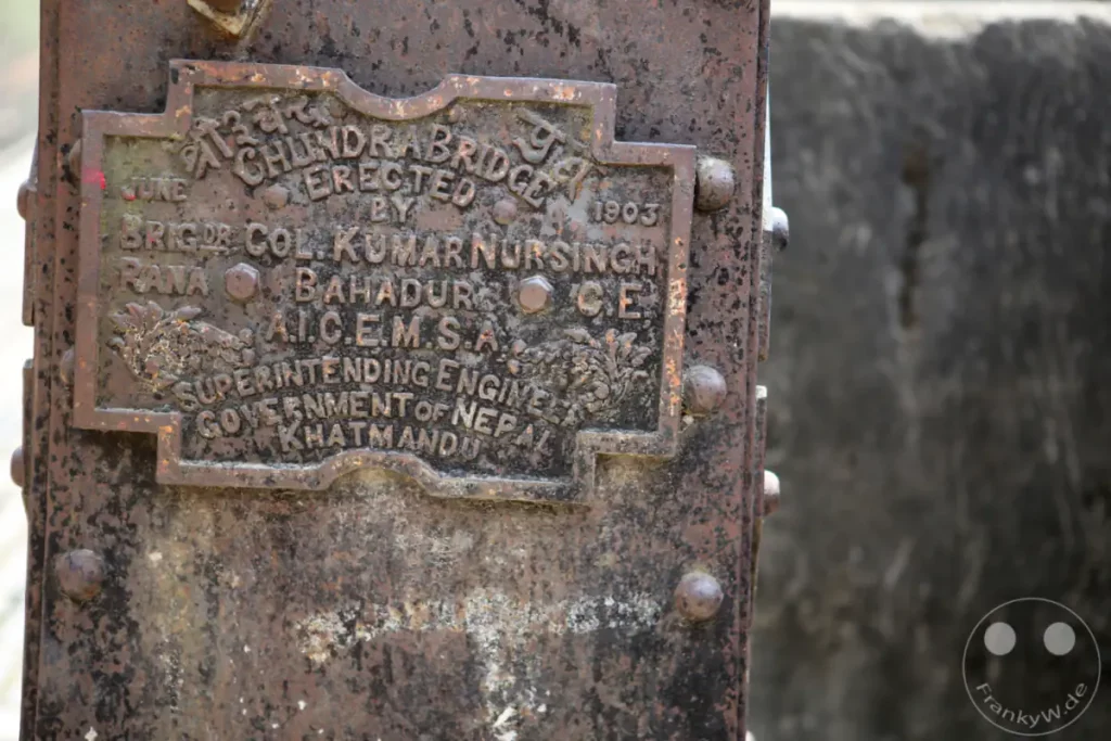 Eine verwitterte Gedenktafel aus Metall an der Chandra Bridge in Kathmandu, Nepal, dokumentiert deren Erbauungsdatum und die beteiligten Personen. Die Korrosion des Metalls verleiht der Aufnahme eine melancholische und historische Stimmung.