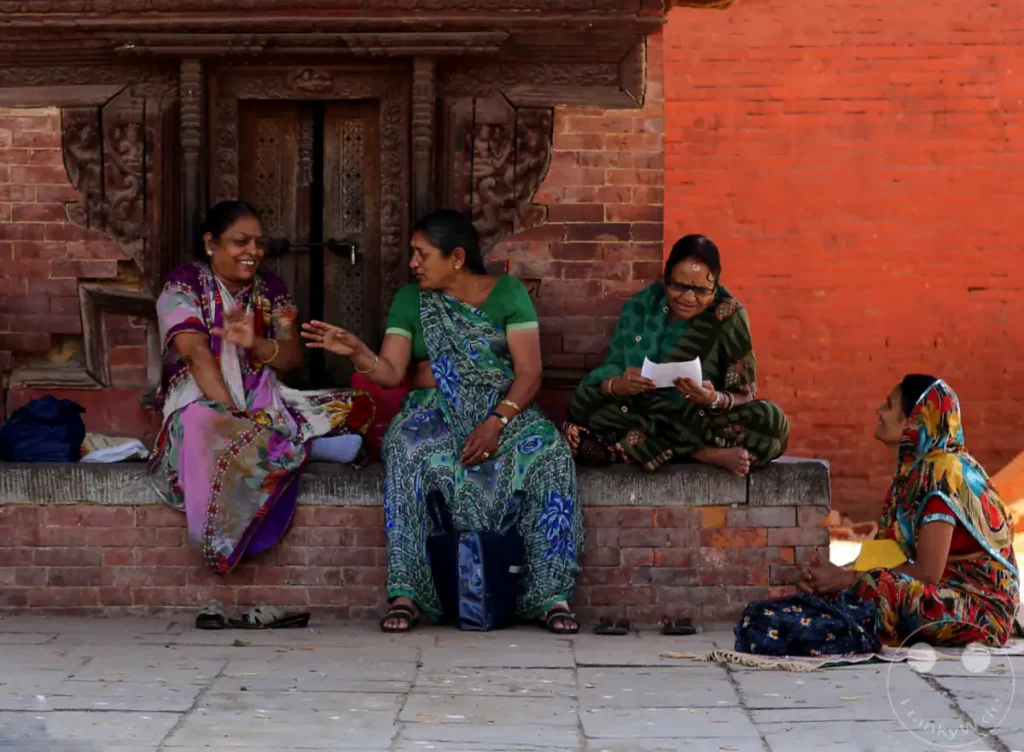 Vier Frauen, gekleidet in Saris, sitzen auf einer Steinmauer vor einem Holztor am Kathmandu Durbar Square. Zwei Frauen unterhalten sich lebhaft, während eine dritte Zeitung liest und die vierte in Gedanken versunken nach oben schaut. Die Szene vermittelt eine Atmosphäre des Alltags und der Gemeinschaft inmitten eines historischen Ortes.