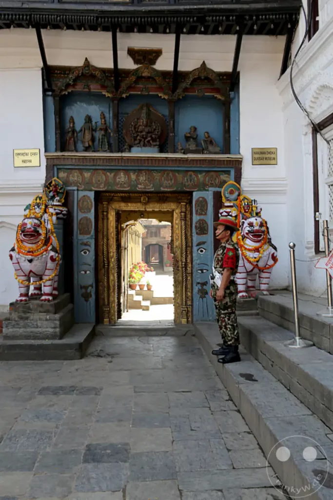 Ein Wachmann in Uniform steht am Eingang des Hanuman Dhoka Durbar Museum am Kathmandu Durbar Square. Vor dem Eingang sind kunstvoll verzierte Löwenstatuen auf Podesten aufgestellt, die eine traditionelle nepalesische Architektur und Kultur widerspiegeln.