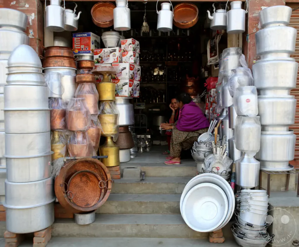 Ein Laden auf dem Kathmandu Durbar Square in Nepal, der eine Vielzahl von Metallwaren verkauft. Auf den Stufen vor dem Laden sind gestapelte Töpfe und Schalen aus Aluminium und Kupfer zu sehen. Zwei Frauen sitzen vor dem Laden und unterhalten sich.