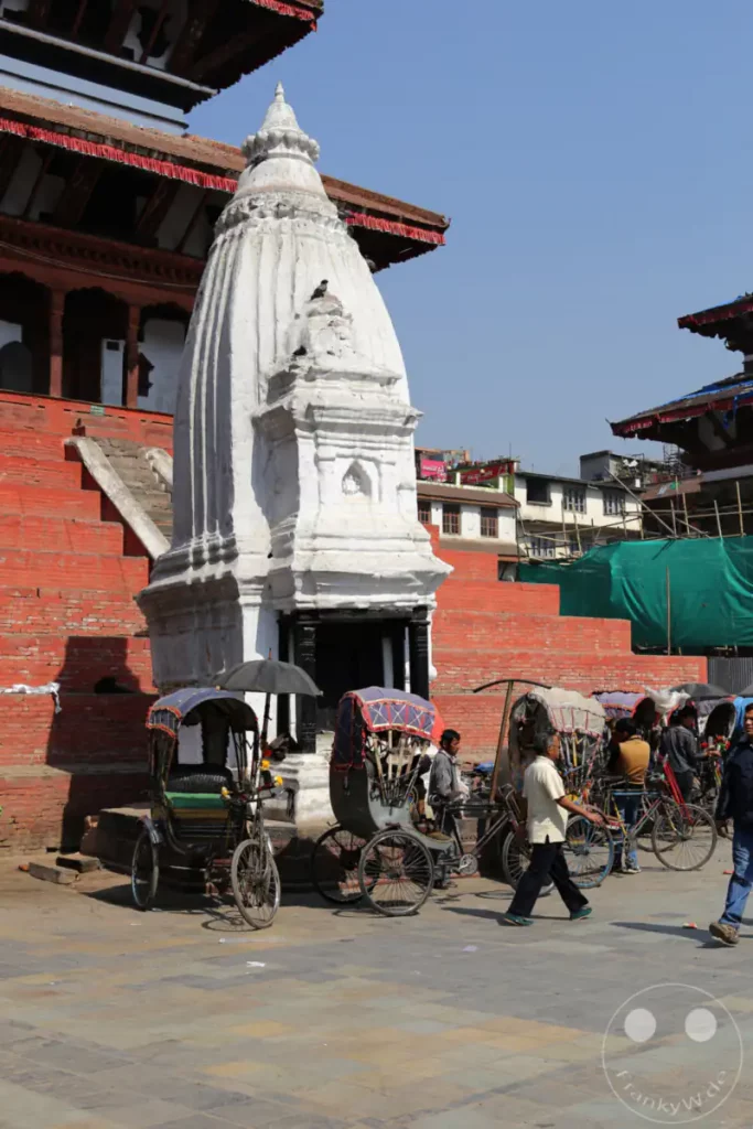 In der lebhaften Kulisse des Kathmandu Durbar Square stehen traditionelle Rikschas bereit, während im Hintergrund ein weißer Tempel mit kunstvollen Schnitzereien aufragt. Die Szene strahlt das geschäftige Alltagsleben und die kulturelle Tiefe des historischen Platzes aus.