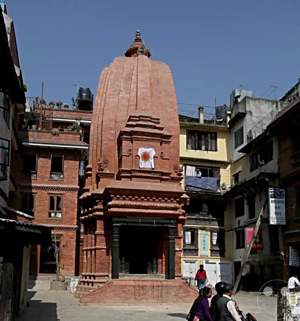 Ein detailreicher Ziegel-Tempel dominiert die Szene auf dem Kathmandu Durbar Square. Umgeben von traditionellen Gebäuden und belebter Atmosphäre zeigt das Bild die kulturelle Vielfalt und historische Architektur Nepals. Menschen gehen ihren täglichen Geschäften nach, während der altehrwürdige Tempel im Mittelpunkt steht.