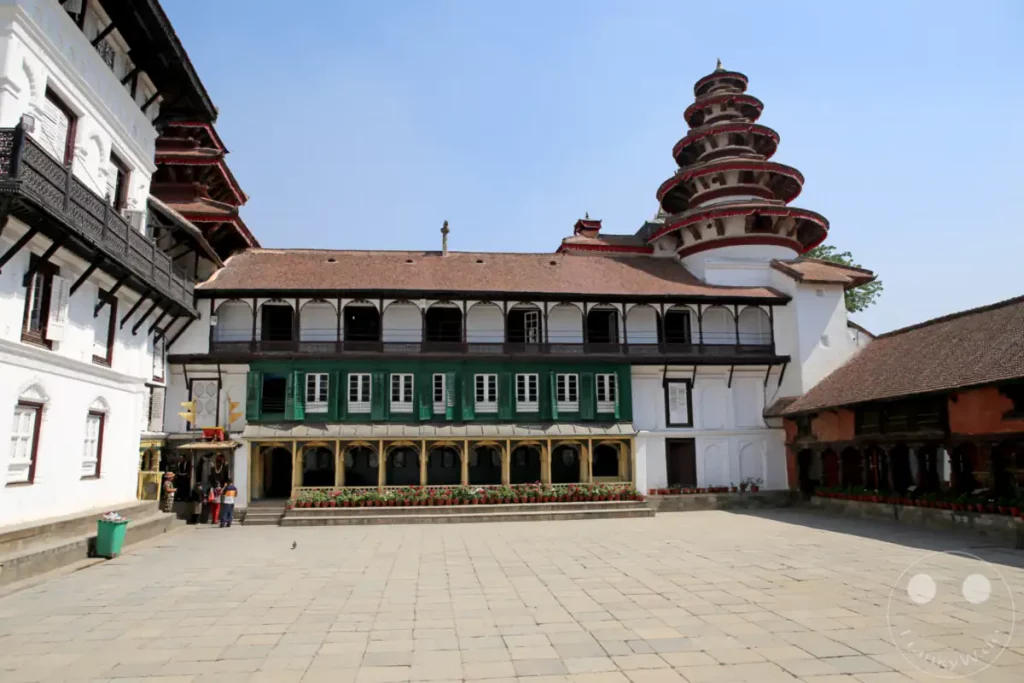 Eine Ansicht des Kathmandu Durbar Square mit seiner reich verzierten nepalischen Architektur. Die Gebäude zeichnen sich durch ihre detailreichen Schnitzereien und traditionellen Pagodendächer aus, die vor einem klaren blauen Himmel stehen. Der Platz ist mit Steinplatten ausgelegt und wirkt ruhig und majestätisch.