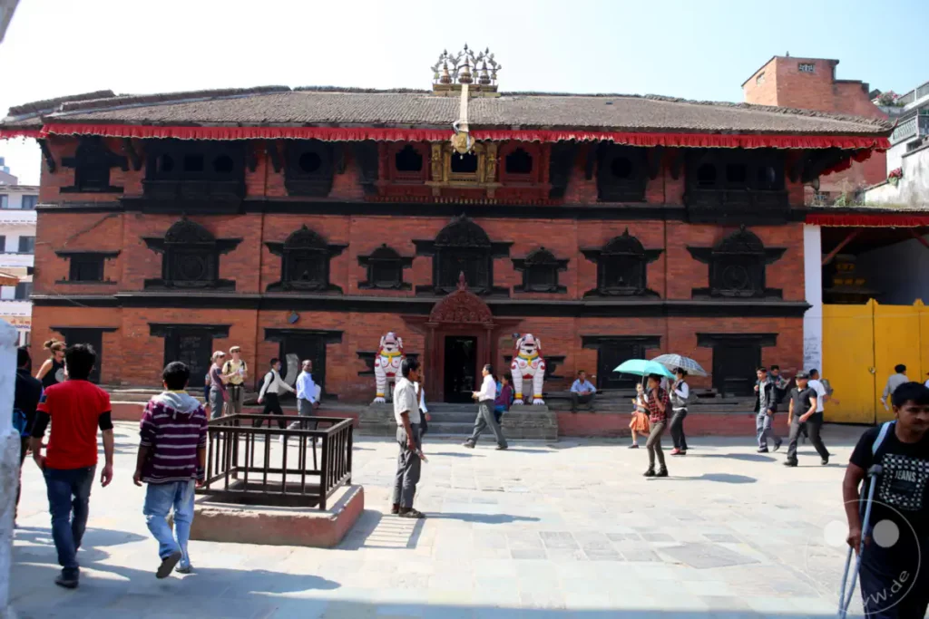 Der Kathmandu Durbar Square in Nepal zeigt eine traditionelle Tempelarchitektur aus Ziegeln, geschmückt mit roten Stoffbahnen und kunstvollen Schnitzereien. Menschen unterschiedlichen Alters bewegen sich auf dem gepflasterten Platz, einige mit Regenschirmen, während die Sonne hell scheint.