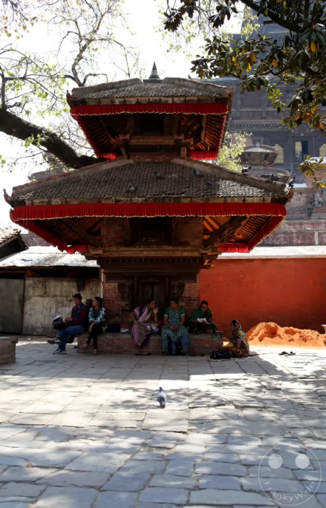 Eine Gruppe von Menschen sitzt auf einer Mauer vor einem mehrstöckigen Tempel am Kathmandu Durbar Square. Das Bauwerk ist mit roten Stoffbahnen geschmückt, und Tauben sind auf dem Dach zu sehen. Der Platz ist mit Steinplatten gepflastert und von Bäumen beschattet.
