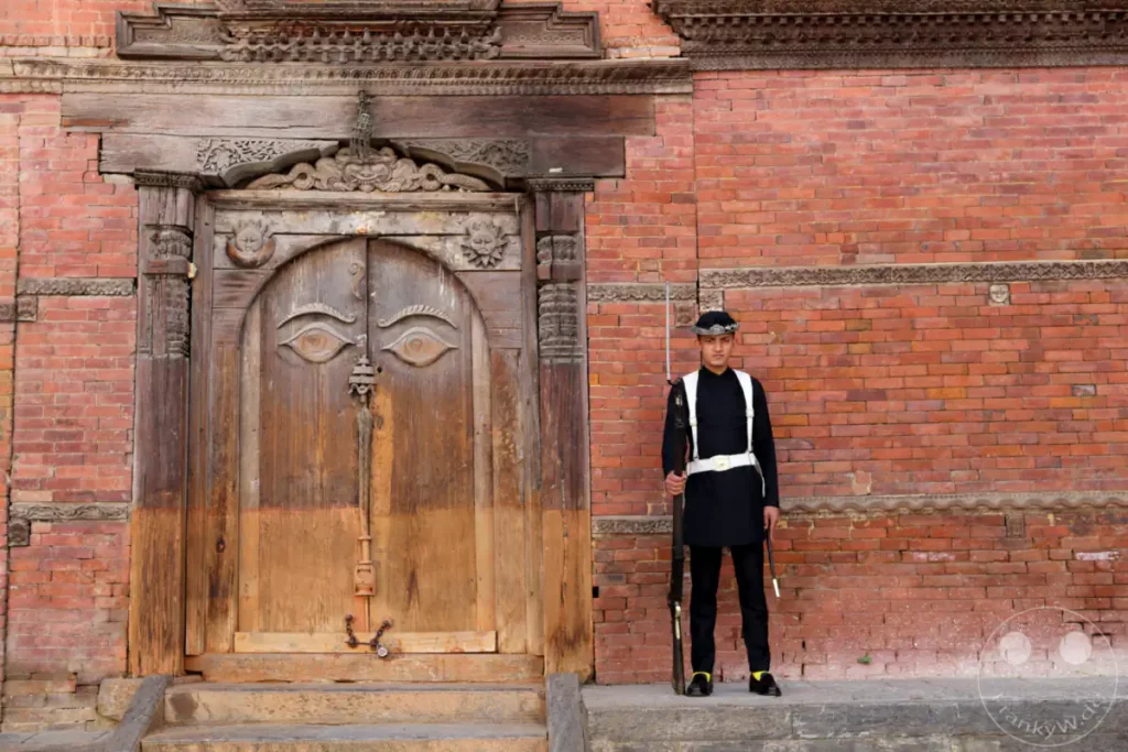 Ein Wachposten in traditioneller Uniform steht vor einer kunstvoll geschnitzten Holztür am Kathmandu Durbar Square. Die Szene kombiniert historische Architektur mit der Präsenz eines modernen Wächters und vermittelt eine Atmosphäre von Ehrfurcht und Beständigkeit.
