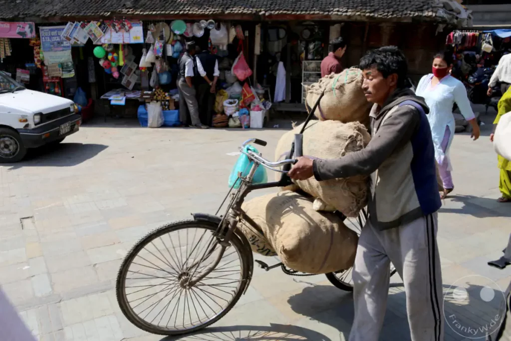 Ein Händler schiebt ein mit Säcken beladenes Fahrrad durch die belebte Gasse des Kathmandu Durbar Square. Die Szene zeigt das geschäftige Treiben auf dem Markt und den Transport von Waren mit einfachen Mitteln.