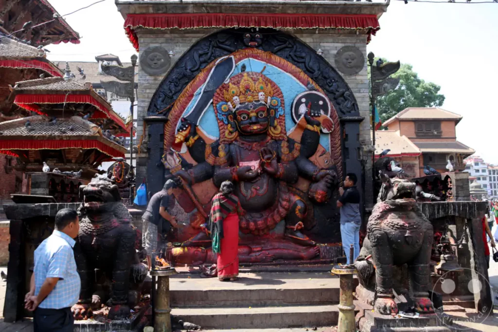 Eine farbenprächtige, detailreiche Statue einer hinduistischen Gottheit dominiert den Platz am Kathmandu Durbar Square. Mehrere Personen und eine friedliche Taubenschar sind auf und um die Kultstätte versammelt, was eine ruhige und spirituelle Atmosphäre erzeugt.