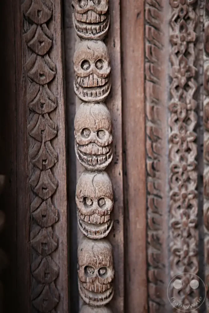 Eine detaillierte Nahaufnahme einer kunstvollen Holzschnitzerei, die eine Reihe von grinsenden Totenschädeln zeigt. Die Verzierungen, wahrscheinlich Teil eines alten Torbogens am Kathmandu Durbar Square, zeugen von traditioneller Handwerkskunst und verleihen dem Bild eine düstere, aber faszinierende Atmosphäre.
