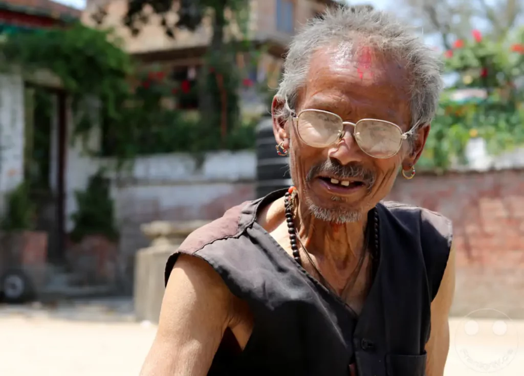 Ein älterer Mann mit Brille und traditioneller Bemalung auf der Stirn blickt in die Kamera. Er trägt mehrere Halsketten und Ohrringe, was seine spirituelle oder kulturelle Identität unterstreicht. Die Aufnahme in Kathmandu fängt eine Momentaufnahme seines Lebens und seiner Persönlichkeit ein, vermittelt eine ruhige und nachdenkliche Stimmung.