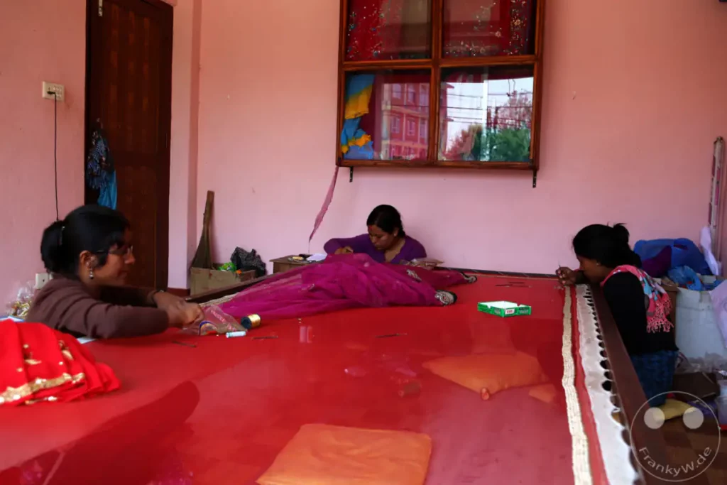 Drei Frauen in Kathmandu sitzen an einem großen, roten Arbeitstisch und besticken kunstvoll einen Saristoff. Mit Nadel und Faden arbeiten sie akribisch an den feinen Verzierungen des Kleidungsstücks. Die Szene strahlt eine Atmosphäre konzentrierter Handwerkskunst und traditioneller Textilherstellung aus.