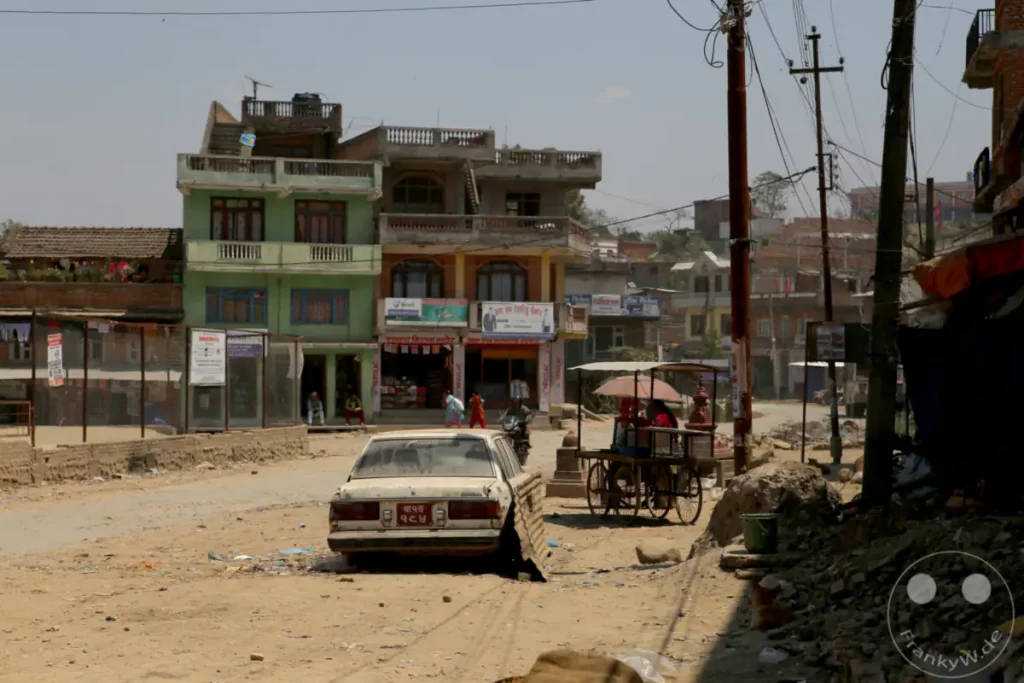 In Kathmandu herrscht geschäftiges Treiben auf einer staubigen Straße, gesäumt von Geschäften und Wohnhäusern. Ein alternder Kombi parkt am Straßenrand, während ein Karren mit einer Marktfrau davor steht. Die Szene vermittelt einen Eindruck von Alltagsleben in einer belebten nepalesischen Stadt.