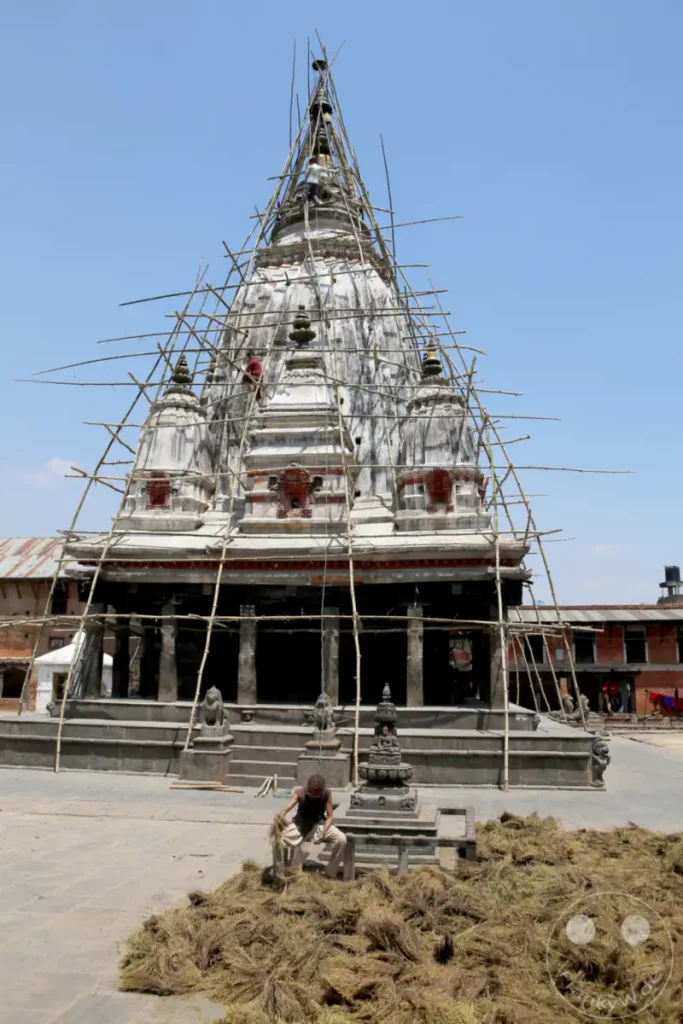 Ein Arbeiter führt Reparaturen an einem traditionellen Tempel in Kathmandu durch, der von einem Bambusgerüst umgeben ist. Vor dem Tempel liegen große Mengen getrockneter Pflanzenmaterialien, möglicherweise für Bauzwecke oder Rituale. Die Szene vermittelt ein Gefühl von fortlaufender Arbeit und kultureller Pflege unter blauem Himmel.