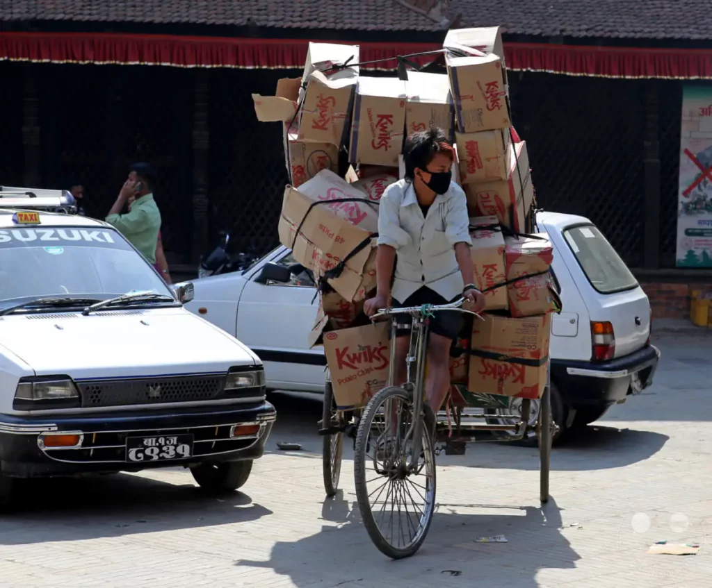 Ein beladener Mann transportiert auf einem Fahrrad mit Anhänger Kisten mit Waren in Kathmandu, Nepal. Mehrere Autos und Gebäude im Hintergrund deuten auf eine städtische Umgebung hin. Die Szene vermittelt eine geschäftige Atmosphäre des Handels und der täglichen Arbeit.