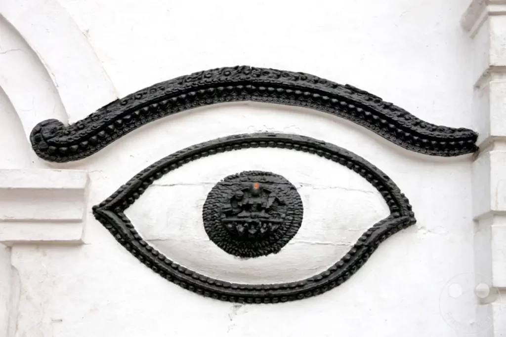 Detailaufnahme eines kunstvollen Augen-Symbols am Pashupatinath-Tempel in Kathmandu, Nepal. Die schwarze, verzierte Darstellung zeigt im Zentrum eine kleine Gottheit und symbolisiert spirituelle Wachsamkeit. Die strahlend weiße Wand bildet einen starken Kontrast zu dem dunklen Kunstwerk.