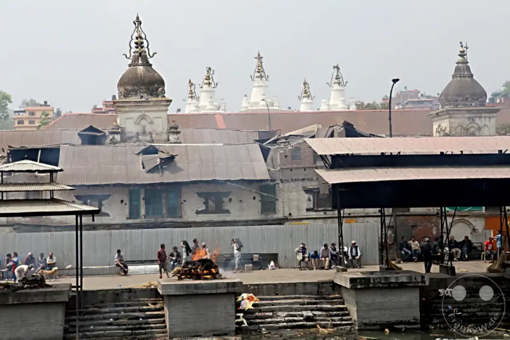 Ein Leichenbrand findet am Ufer des Bagmati-Flusses statt, einem heiligen Fluss in Kathmandu. Menschen versammeln sich, um den Verstorbenen die letzte Ehre zu erweisen, während andere auf den Stufen sitzen und zusehen. Die Szene vermittelt eine Atmosphäre von Andacht und Ritual, die tief in der hinduistischen Kultur verwurzelt ist.