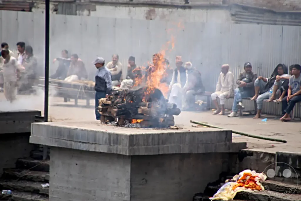 Am Pashupatinath-Tempel in Kathmandu finden rituelle Feuerbestattungen statt, wobei Holzscheite zu Ehren der Verstorbenen angezündet werden. Der Rauch und die Flammen erzeugen eine ernste und nachdenkliche Atmosphäre, während Angehörige und Beobachter im Hintergrund anwesend sind.