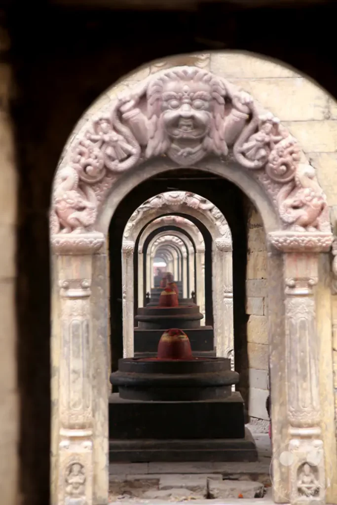 Ein Blick durch eine Reihe von Torbögen im Pashupatinath-Tempel in Kathmandu, Nepal. In der Mitte jedes Bogens sind mehrere Lingams aufgereiht, die auf eine spirituelle Atmosphäre hinweisen.