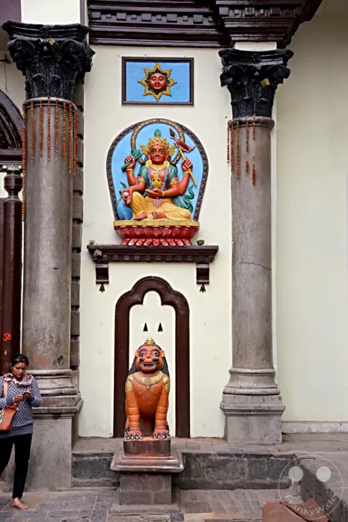 Dieses Bild zeigt hinduistische Skulpturen vor dem Pashupatinath-Tempel in Kathmandu, Nepal. Die detailreichen Darstellungen von Gottheiten und Fabelwesen spiegeln die reiche religiöse und kulturelle Tradition des Ortes wider. Eine Frau im Vordergrund betrachtet ihr Handy, was einen Kontrast zwischen Tradition und Moderne darstellt.