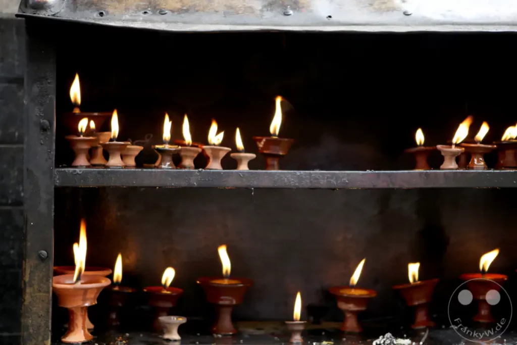 Mehrere kleine Tonkerzen mit brennenden Flammen stehen auf zwei Regalen in einem dunklen Schrein an der Swayambhunath-Stupa. Das warme Licht der Kerzen erzeugt eine andächtige und friedliche Atmosphäre.