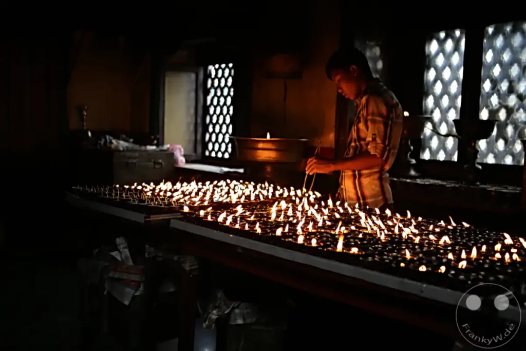 Ein junger Mann zündet in der Swayambhunath-Stupa bei Kathmandu eine Vielzahl von Öllampen an. Die Szene ist in warmes Licht getaucht und strahlt eine ruhige, andächtige Atmosphäre aus, die durch die zahlreichen Flammen verstärkt wird.