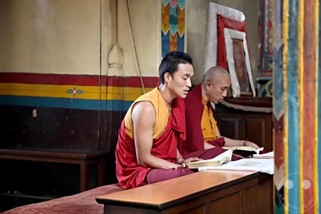 Zwei buddhistische Mönche in traditionellen Gewändern sitzen auf dem Boden und studieren heilige Texte in einem Tempel an der Swayambhunath-Stupa in Kathmandu. Die Szene strahlt eine Atmosphäre der Hingabe und des Lernens aus, mit farbenfrohen Wandmalereien und Holzelementen im Hintergrund.