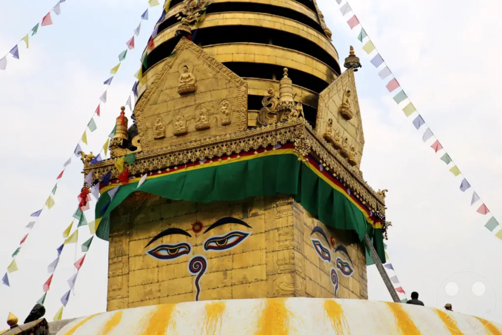 Die goldene Spitze der Swayambhunath-Stupa in Kathmandu, Nepal, ist mit bunten Gebetsfahnen geschmückt. Die charakteristischen Buddha-Augen auf der Stupa blicken in die Ferne, während wenige Menschen die Anlage besuchen. Die friedliche Atmosphäre der Stätte wird durch die leuchtenden Farben und die spirituelle Architektur verstärkt.