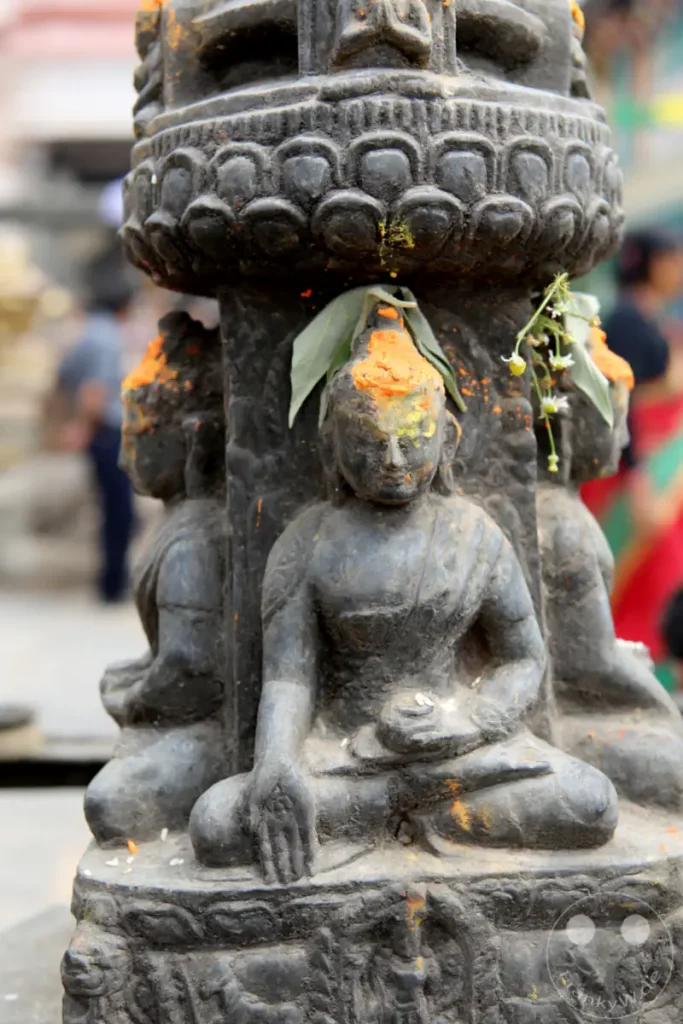 Mehrere Buddha-Statuen sind an einem steinernen Pfeiler in der Nähe der Swayambhunath-Stupa in Kathmandu, Nepal, aufgereiht. Die Statuen sind mit orangefarbenem Pulver und Blumen geschmückt, was auf eine religiöse oder zeremonielle Nutzung hinweist. Die Szene vermittelt ein Gefühl von Spiritualität und Tradition.