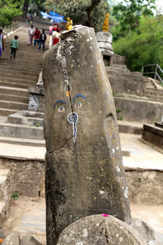 Ein stehender Fels mit bemalten buddhistischen Augen steht im Vordergrund, während im Hintergrund eine Treppe zur Swayambhunath-Stupa in Kathmandu führt. Die Szene vermittelt eine Atmosphäre der Spiritualität und kulturellen Bedeutung inmitten der natürlichen Umgebung.