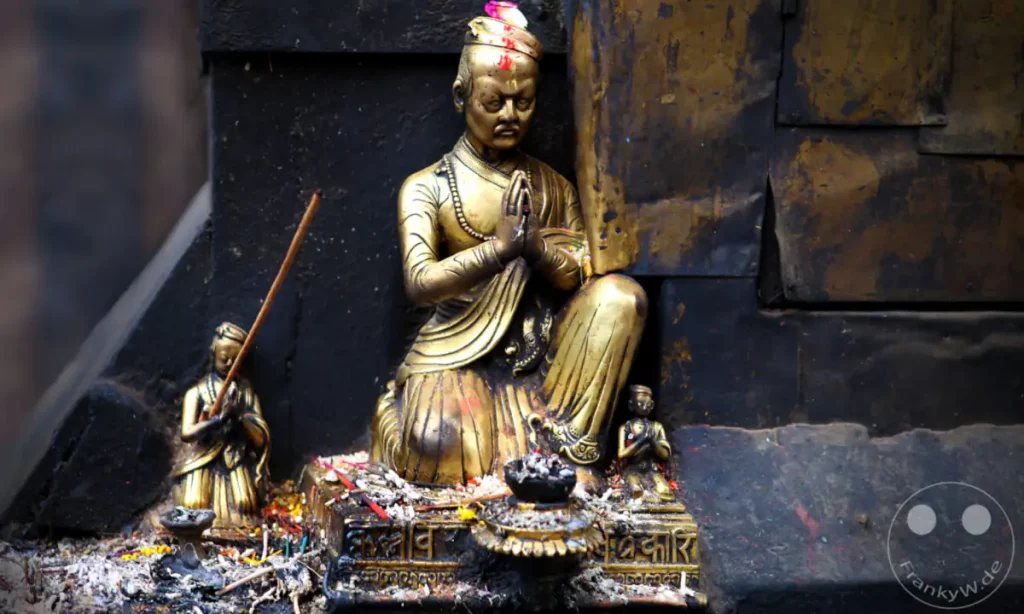 Eine detailreiche Aufnahme von goldenen buddhistischen Gebetsfiguren in der Nähe der Swayambhunath-Stupa in Kathmandu. Die Figuren sind mit Blumen und Räucherstäbchen geschmückt, was eine spirituelle Atmosphäre erzeugt.