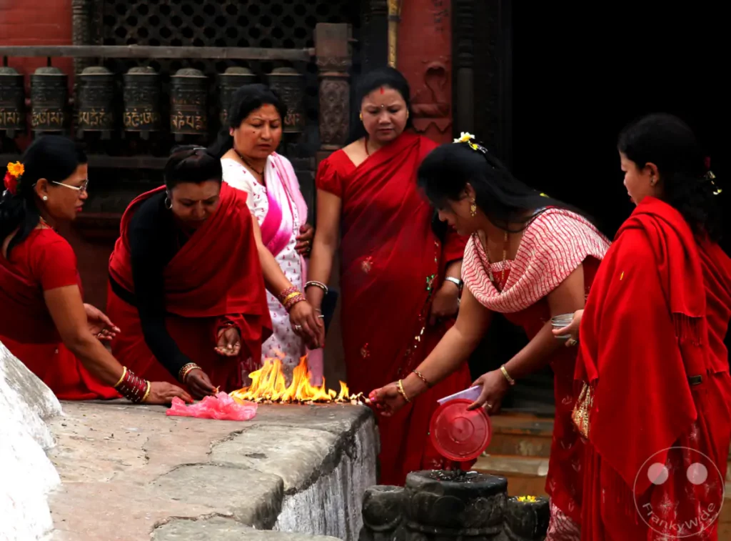 Mehrere Frauen in traditionellen roten Saris versammeln sich um ein heiliges Feuer an der Swayambhunath-Stupa in Kathmandu, Nepal. Sie beteiligen sich an einem religiösen Ritual und werfen Gaben ins Feuer, was eine Atmosphäre der Hingabe und Gemeinschaft erzeugt.