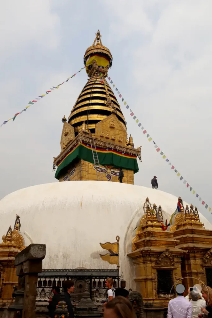 Die monumentale weiße Kuppel des Swayambhunath-Stupa dominiert den Vordergrund, geschmückt mit goldenen Verzierungen und buddhistischen Augen. Über der Kuppel erhebt sich der goldene Turm des Stupa, der von bunten Gebetsfahnen umrahmt wird, die im Wind flattern. Die Szene strahlt eine tiefe spirituelle Atmosphäre aus, verstärkt durch die Anwesenheit von Pilgern und die majestätische Architektur.