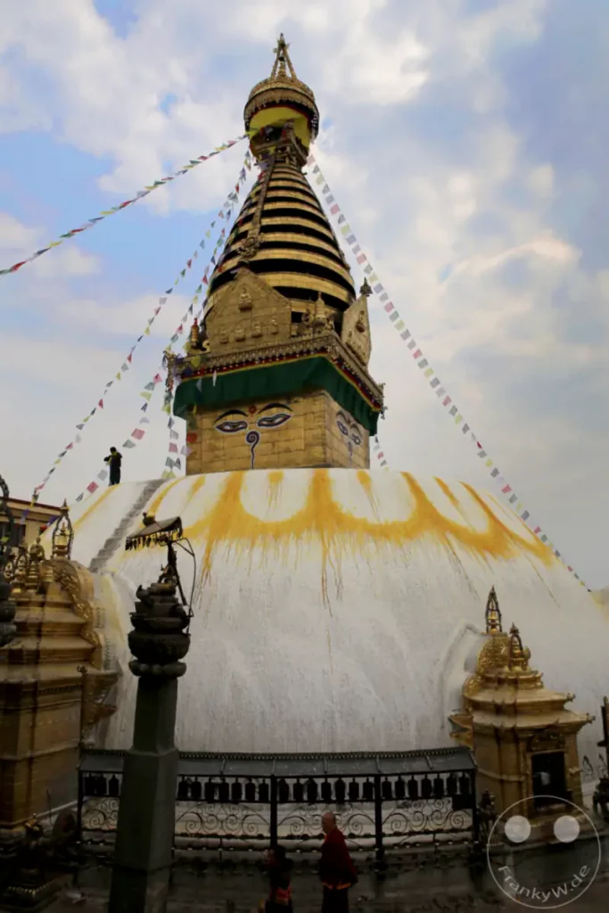 Die monumentale weiße Kuppel des Swayambhunath-Stupa dominiert den Vordergrund, geschmückt mit goldenen Verzierungen und buddhistischen Augen. Über der Kuppel erhebt sich der goldene Turm des Stupa, der von bunten Gebetsfahnen umrahmt wird, die im Wind flattern. Die Szene strahlt eine tiefe spirituelle Atmosphäre aus, verstärkt durch die Anwesenheit von Pilgern und die majestätische Architektur.